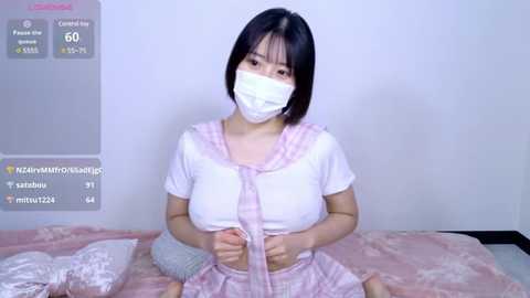 Media: A young Asian woman with short black hair and fair skin, wearing a pink and white sailor schoolgirl outfit, mask, and gloves, kneeling on a bed with pink sheets in a minimalist room.