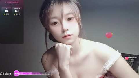 Media: Video of a young East Asian woman with fair skin, dark hair, and a slender build, wearing a white off-shoulder dress, resting her chin on her hand, with a grey background and digital overlay.