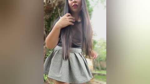 Media: Video of an Asian woman with long, straight black hair, wearing a black sleeveless top and a checkered skirt, standing outdoors by a green, blurred background.