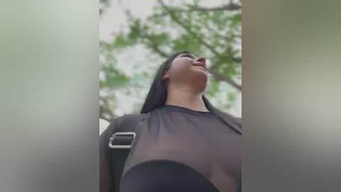 Media: A video of a young woman with medium-length dark hair, wearing a sheer black top, standing in a lush, green forest. The background features blurred tree branches and foliage.