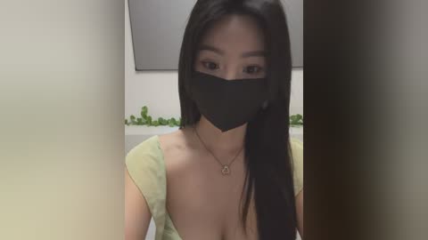 Media: Video of an East Asian woman with long black hair, wearing a black face mask, green top, and a necklace, indoors with green plants in the background.