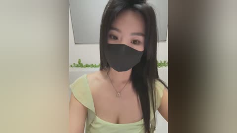 Media: Video of an Asian woman with long black hair, wearing a black face mask and a light green off-shoulder top, indoors with a white wall and green potted plants in the background.