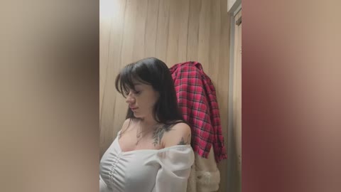 Media: A video of a pale-skinned woman with dark hair, wearing a white off-the-shoulder top, leaning against a wooden door with a red plaid shirt draped over her shoulders.