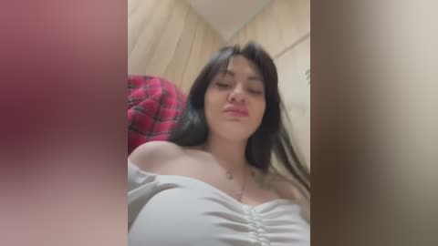 Media: Video of a young woman with long dark hair, fair skin, and large breasts, wearing a white off-shoulder top. Background shows a red plaid blanket and light wood-paneled wall.