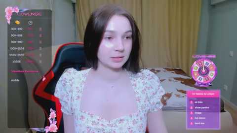 Video of a young woman with straight brown hair, wearing a floral top, sitting in a gaming chair. Background features a bed, a cow print blanket, and a fortune-telling wheel.