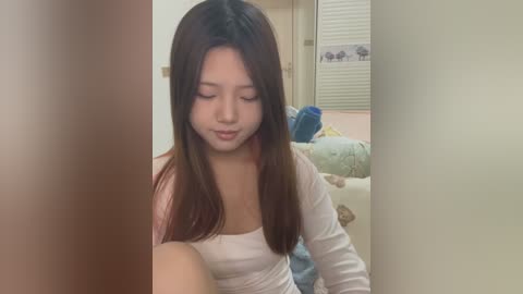 Media: Video of a young Asian woman with straight, shoulder-length brown hair, wearing a white long-sleeved top, sitting on a bed in a dimly lit room with closed blinds and a blue blanket.