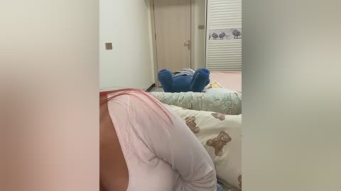 Media: Video of a bedroom with a person lying on a bed, covered by a light pink and brown patterned blanket, wearing white pants. A blue pillow and a beige blanket are visible in the background.