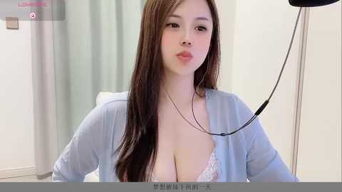 Media: A video of a young East Asian woman with long, straight brown hair, wearing a light blue cardigan and revealing a pink lace bra, sitting in a medical exam room with beige curtains.