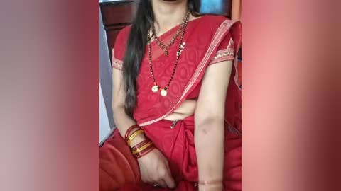 Media: Video of a South Asian woman in a red sari, adorned with gold jewelry, seated indoors. Her dark hair is long and loose, and she wears multiple bangles on her wrist.