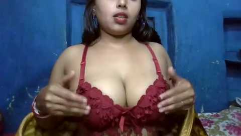 Media: Video of a young woman with medium brown skin and long black hair, wearing a red lace bra, pressing her breasts together. She stands in a dimly lit room with blue walls, and a bed with floral sheets in the background.