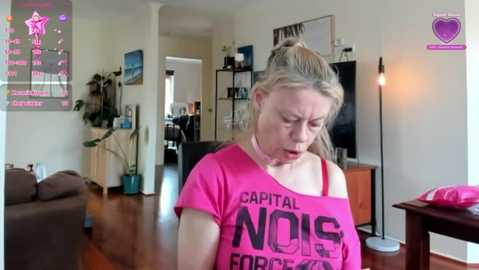 Media: A video of a fair-skinned, blonde-haired woman in a pink \"Capital NDIS\" t-shirt, looking contemplative, in a bright, modern living room with a large TV and bookshelf in the background.