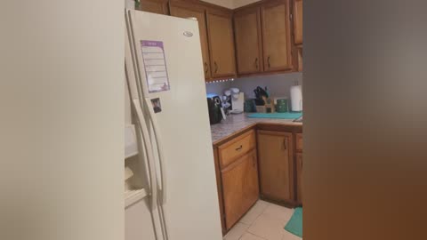 Media: Video of a small, outdated kitchen with wooden cabinets, a white refrigerator, and a cluttered counter with appliances and utensils. The image is slightly blurred.