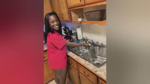 Media: A video of a smiling Black woman with long black hair, wearing a red T-shirt and beige shorts, washing dishes in a wooden kitchen.