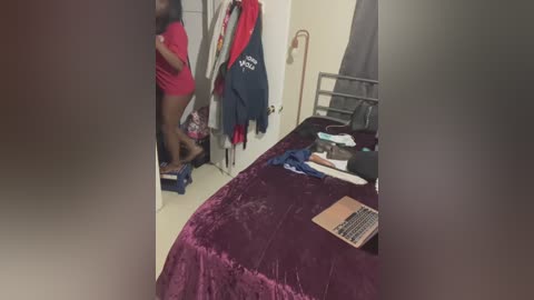 Media: Video of a messy bedroom with a large, unmade bed covered in a maroon comforter. Clothes, including a red shirt, are scattered around. A mirror reflects a person in red pants and a black top.