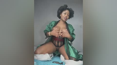 Media: Video of a Black woman with dark skin, wearing a green satin robe, lace lingerie, and black hair styled in a bouffant, kneeling on a blue bedspread, hands covering her breasts, indoors.