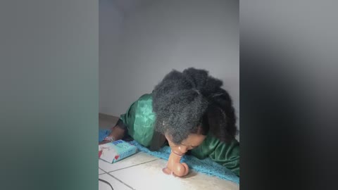 Video of a young Black child with a curly afro, lying on a tiled floor in a dimly lit room, wearing a green shirt. The child is reaching towards a blue blanket.