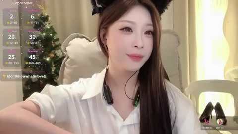 Media: Video of an East Asian woman with long brown hair, wearing a white blouse, black headphones, and cat ears. She's seated in a cozy room with a Christmas tree, calendar overlay, and high heels on a table.