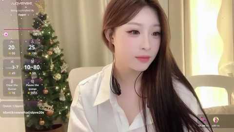 Media: Video of an Asian woman with long brown hair, wearing a white blouse, sitting next to a decorated Christmas tree in a cozy room.