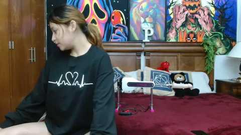 Video of a young woman with light skin and straight, light brown hair, wearing a black sweatshirt with a white heart symbol. She sits on a red bed with a wooden headboard and colorful posters.