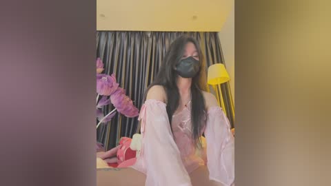 Media: Video of a woman with long black hair and light skin, wearing a pink off-shoulder top, black face mask, and pink headband, standing in a dimly lit room with dark curtains and a yellow lamp.