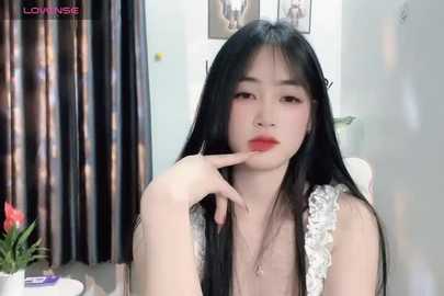 Media: Video of an Asian woman with long black hair, wearing a white lace top, sitting indoors, touching her lips, surrounded by curtains and a potted plant.