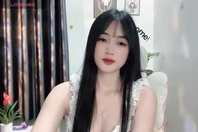 Media: A video of a young Asian woman with long black hair, fair skin, wearing a white lace top, sitting in a room with dark curtains and plants.