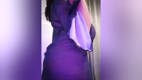 Media: Video of a person with dark skin, partially obscured, holding a white towel, standing in a dimly lit room with purple lighting, creating a soft, sensual atmosphere.