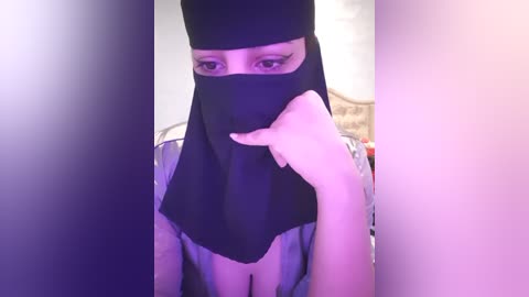 Media: A video of a woman with light skin, wearing a black hijab, sitting in a room with purple and white lighting. She has a delicate, feminine face and is looking directly at the camera.