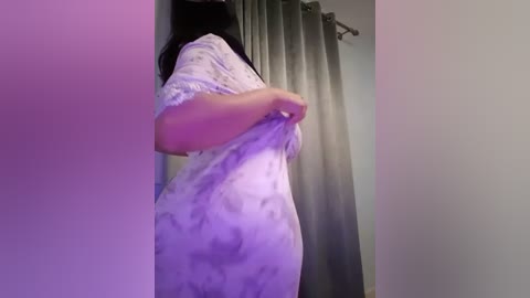 Media: Video of a woman in a white, tie-dye dress, holding the fabric, facing a gray curtain in a dimly lit room.