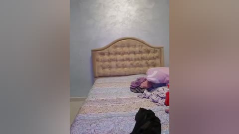 Media: Video of a messy bedroom with a light blue wall, a beige tufted headboard, and a bed covered in pastel-patterned sheets, featuring disheveled bedding and a black garment.