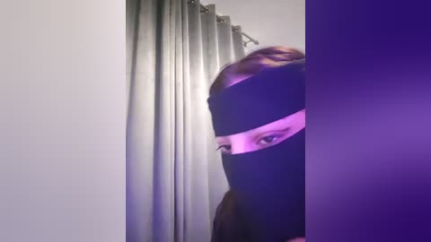 Media: Video of a person with a black mask covering their face, peering from behind a gray curtain, casting a purple light on their features.