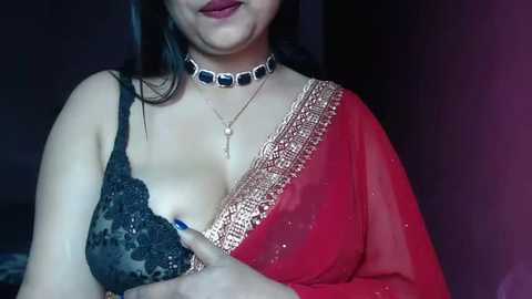 Media: Video of a light-skinned woman with long dark hair, wearing a black lace bra and a red saree with gold embroidery. She has a silver choker with blue stones and a pendant necklace. The background is dark.