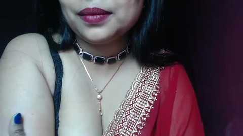 Media: Video of a woman with medium skin tone, dark hair, and full lips wearing a red saree with gold embroidery, a black bra, and a choker necklace. Background is dark.