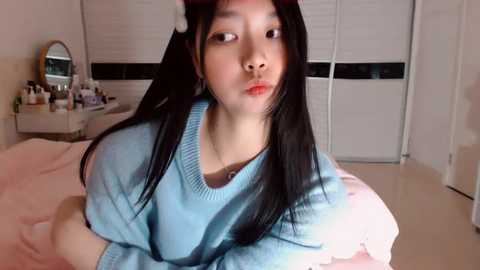 Media: Video of an Asian woman with long black hair, wearing a blue sweater, lying on a pink bed in a brightly lit bedroom with white walls and a vanity table.