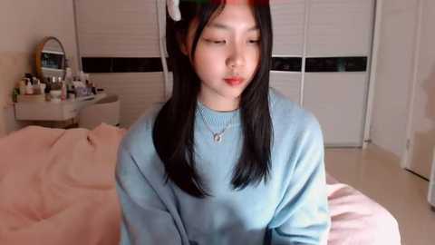 Media: A video of an Asian woman with straight black hair and fair skin, wearing a light blue sweater and red lipstick, sitting on a bed in a dimly lit, cozy bedroom with white walls and a vanity table.