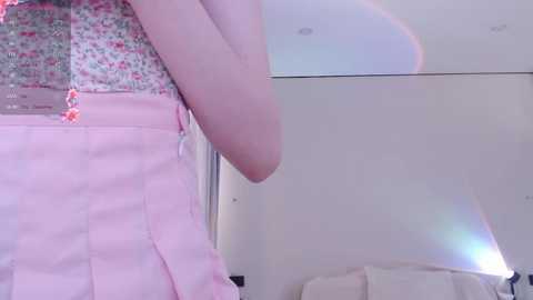 Media: A video of a pale-skinned woman wearing a light pink pleated skirt with a floral print and a white top, standing in a brightly lit bedroom with a reflective mirror and a glowing lamp.