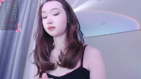 Media: Video of a young Asian woman with fair skin, long brown hair, and closed eyes, wearing a black spaghetti-strap top. Background features a white ceiling and a gray curtain with pink floral patterns.