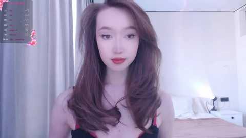 Media: Video of a young, slender, fair-skinned woman with long, wavy brown hair, wearing red lipstick and black lingerie, in a modern, minimalist bedroom with white walls, bed, and curtains.