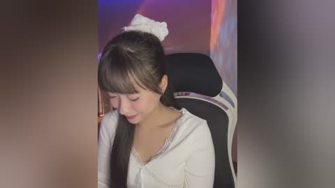 Media: A video of a young East Asian woman with long black hair, wearing a white top, seated in a black and white gaming chair, with a blurred background.