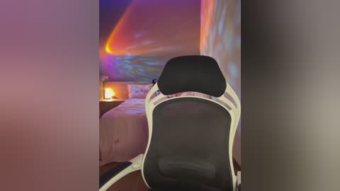 Media: A video shows a black gaming chair with white accents and a white pillow in a dimly lit bedroom. The background features a bed, a lit candle on a nightstand, and a wall adorned with colorful, abstract light patterns.