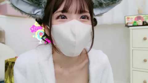 Media: Video of an East Asian woman wearing a white face mask and a furry rabbit headband, with a soft, white robe. Background includes a white dresser and a colorful, abstract painting.