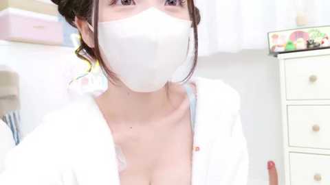 Media: Video of a young woman with light skin, dark hair, and a white mask, wearing a white robe, revealing cleavage. Background features a white dresser with colorful objects and a pink bed.