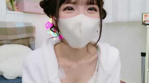 Media: Video of an East Asian woman with fair skin, wearing a white surgical mask, pink earrings, and a white robe, in a cozy, bright room with a plaid couch and white curtains.