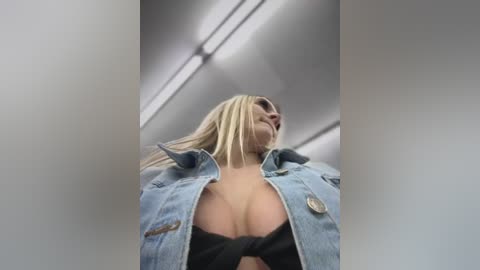 Media: Video of a blonde woman with long hair, wearing a light blue denim jacket, black bra, and sunglasses. She is captured from a low angle, emphasizing her cleavage. The background features a modern, white-walled interior with fluorescent lights.