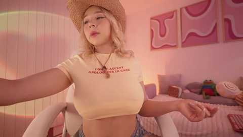 Video of a young woman with fair skin, blonde hair, wearing a beige crop top, a cowboy hat, and denim shorts, standing in a pink bedroom with abstract art on the wall.