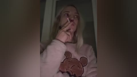 Media: Video of a blonde woman with long hair, wearing a pink bear-themed sweatshirt, holding a phone to her face, indoors with dim lighting.