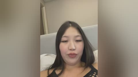 Media: Video of an East Asian woman with long black hair and fair skin, wearing a black lace top, lying on a bed with white pillows, making a kissy face.