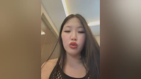 A video of an East Asian woman with long, straight black hair, wearing a black top, puckering her lips in a kiss. The background features a beige wall and a ceiling with recessed lighting.