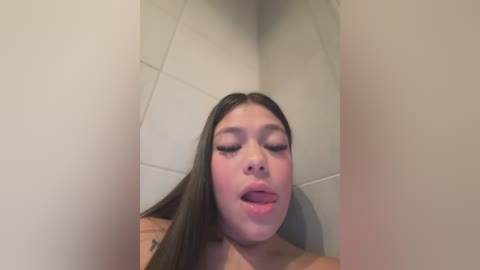 Media: A video of a young Asian woman with long brown hair, fair skin, and closed eyes, sticking out her tongue. She's wearing a beige top and is in a tiled room with beige walls.