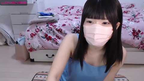 Media: Video of a young East Asian woman with long black hair and light skin, wearing a blue tank top and face mask, kneeling on a colorful rug in a pink-floral bedroom.